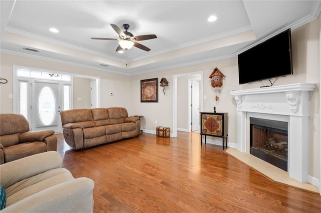 living room with wood-type flooring, ornamental molding, a tray ceiling, ceiling fan, and a fireplace