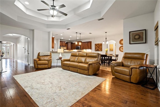 living room with a tray ceiling, dark wood-type flooring, and ceiling fan with notable chandelier