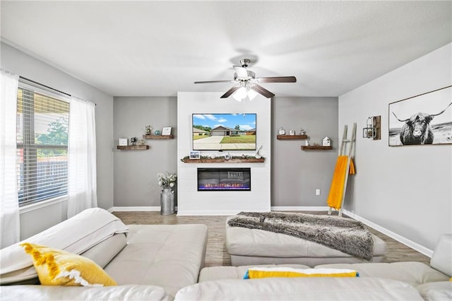 living area featuring wood finished floors, a glass covered fireplace, a ceiling fan, and baseboards