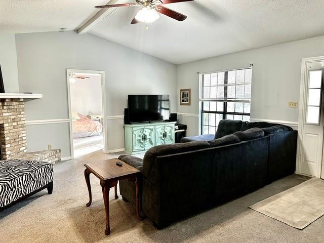 living room featuring a brick fireplace, ceiling fan, lofted ceiling with beams, carpet flooring, and a textured ceiling