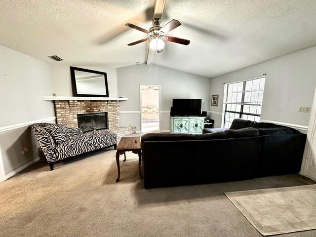 carpeted living area with a ceiling fan, visible vents, lofted ceiling with beams, a textured ceiling, and a brick fireplace