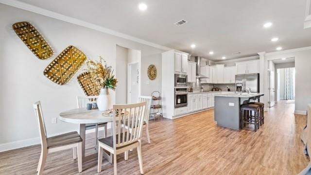 kitchen featuring crown molding, wall chimney range hood, an island with sink, light wood-style flooring, and stainless steel appliances