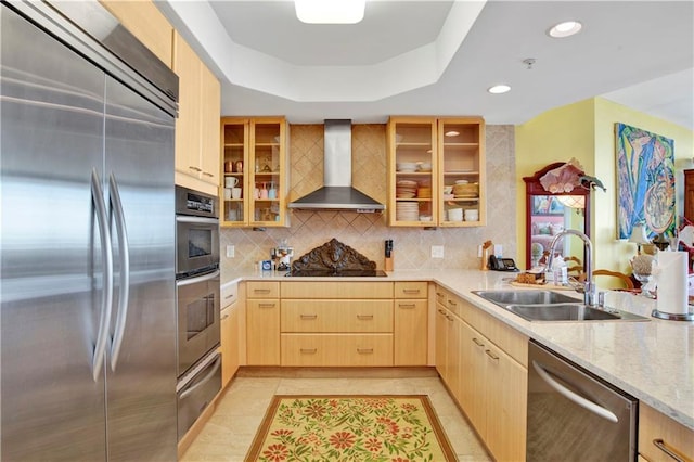 kitchen with wall chimney range hood, a tray ceiling, light brown cabinetry, sink, and stainless steel appliances