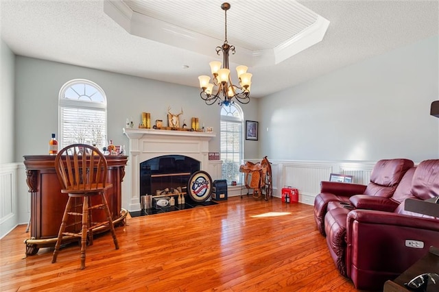 living area featuring hardwood / wood-style flooring, a notable chandelier, a fireplace with flush hearth, wainscoting, and a raised ceiling