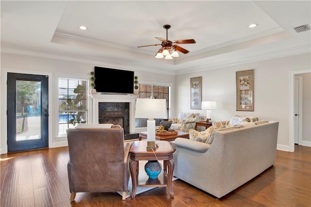 living room with a high end fireplace, a raised ceiling, ornamental molding, dark wood-type flooring, and ceiling fan