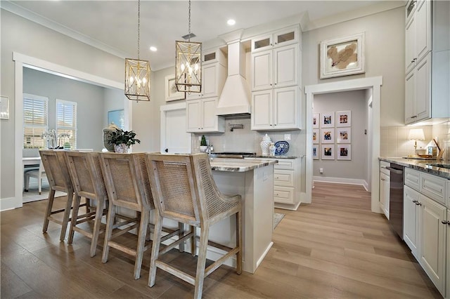 kitchen featuring a kitchen island with sink, white cabinets, hanging light fixtures, light stone countertops, and custom range hood