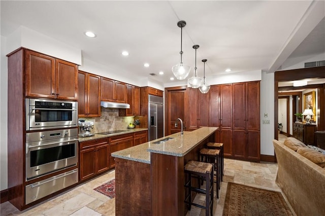 kitchen featuring appliances with stainless steel finishes, light stone counters, sink, decorative light fixtures, and an island with sink