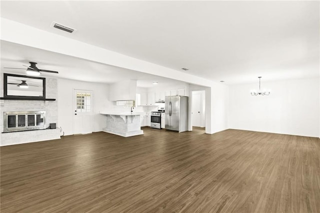 unfurnished living room with ceiling fan with notable chandelier, a brick fireplace, and dark wood-type flooring