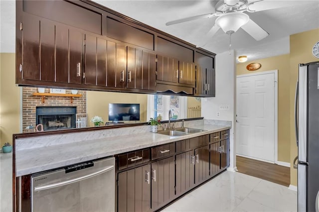 kitchen with a brick fireplace, dark brown cabinets, stainless steel appliances, ceiling fan, and sink