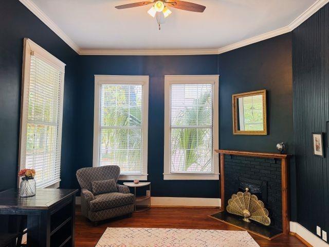 living area with baseboards, ceiling fan, a brick fireplace, crown molding, and wood finished floors