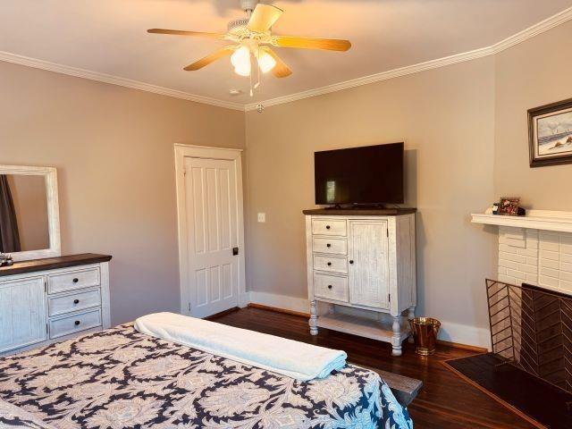 bedroom with a ceiling fan, a brick fireplace, dark wood-style flooring, baseboards, and ornamental molding