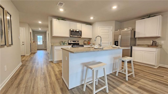 kitchen with an island with sink, white cabinetry, light stone counters, stainless steel appliances, and light hardwood / wood-style flooring