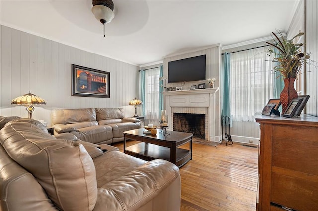 living area featuring light wood finished floors, baseboards, ceiling fan, crown molding, and a brick fireplace