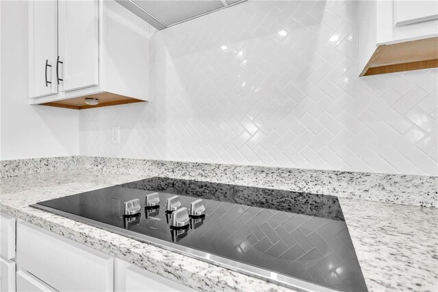 kitchen view of black electric stovetop, white cabinets, decorative backsplash, and light stone countertops