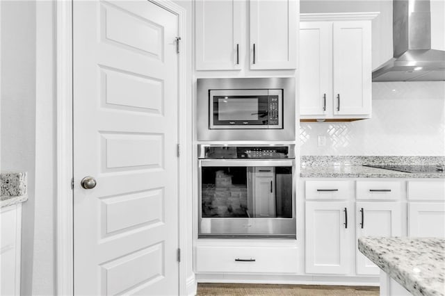 kitchen featuring stainless steel appliances, wall chimney exhaust hood, white cabinetry, decorative backsplash, and light stone counters