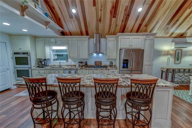 kitchen featuring a breakfast bar, light stone counters, a kitchen island, stainless steel appliances, and wall chimney range hood