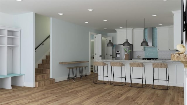 kitchen with wall chimney exhaust hood, stainless steel refrigerator with ice dispenser, a kitchen bar, white cabinets, and light wood-type flooring