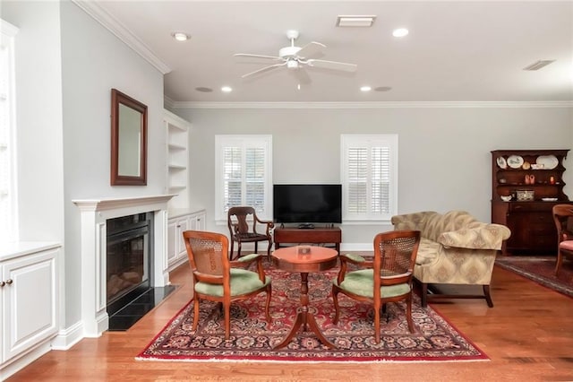 living area featuring visible vents, built in features, a glass covered fireplace, ornamental molding, and light wood-type flooring