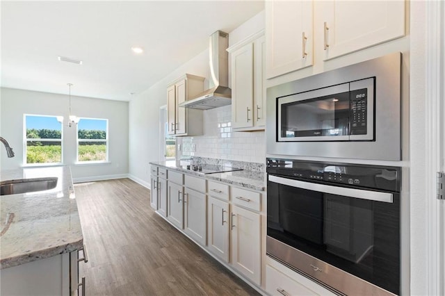 kitchen with light stone counters, pendant lighting, appliances with stainless steel finishes, a sink, and wall chimney range hood