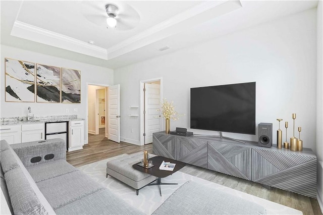 living room with indoor wet bar, crown molding, a raised ceiling, ceiling fan, and wood finished floors