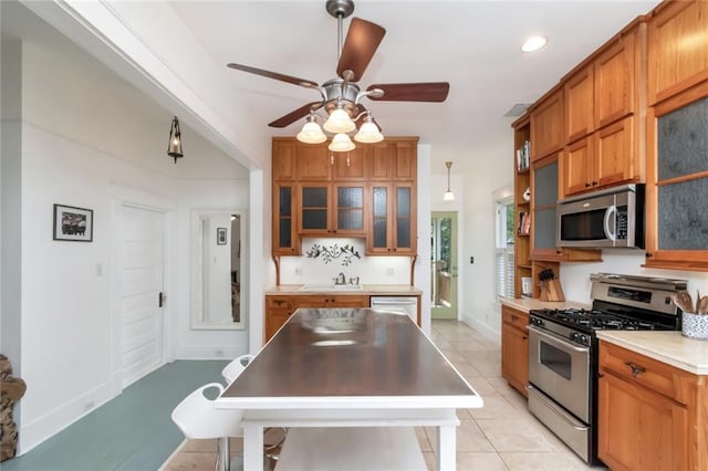 kitchen featuring light tile patterned floors, glass insert cabinets, brown cabinets, a ceiling fan, and appliances with stainless steel finishes
