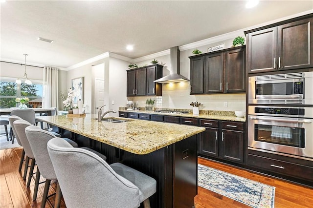 kitchen featuring appliances with stainless steel finishes, wall chimney exhaust hood, a kitchen island with sink, sink, and hardwood / wood-style floors