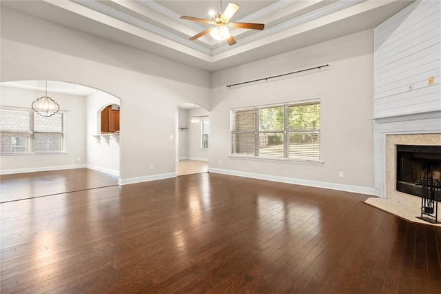 unfurnished living room with dark wood-type flooring, ceiling fan with notable chandelier, crown molding, a premium fireplace, and a tray ceiling