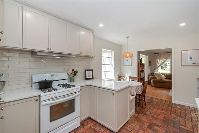 kitchen featuring under cabinet range hood, a peninsula, light countertops, brick floor, and white gas range