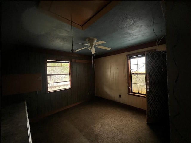 unfurnished room featuring ceiling fan, a healthy amount of sunlight, wooden walls, and dark colored carpet