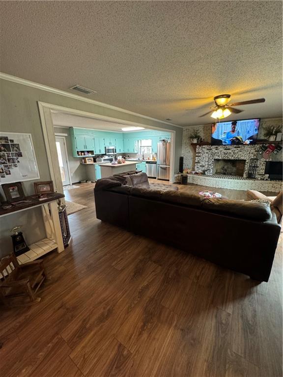 living room featuring crown molding, ceiling fan, dark hardwood / wood-style floors, a textured ceiling, and a stone fireplace