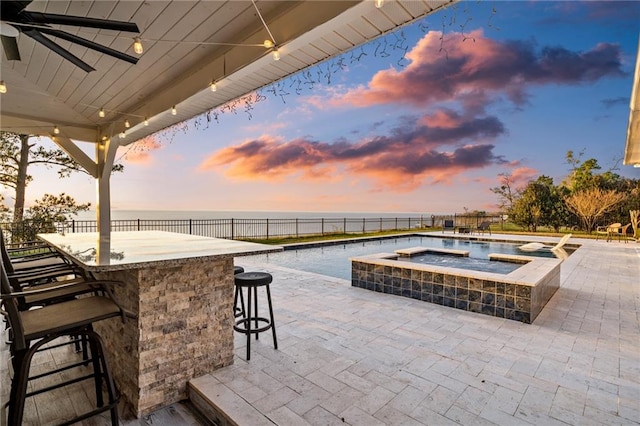 patio terrace at dusk with a water view, an outdoor bar, ceiling fan, and a pool with hot tub