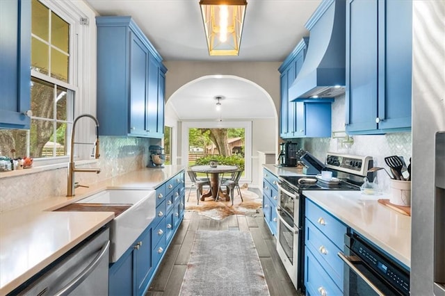 kitchen featuring a sink, stainless steel appliances, blue cabinets, premium range hood, and arched walkways
