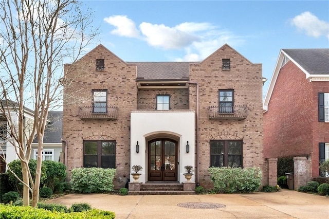 view of front of home with french doors, brick siding, a balcony, and roof with shingles