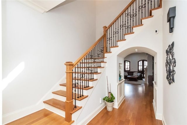 stairway with arched walkways, wood finished floors, a towering ceiling, and baseboards