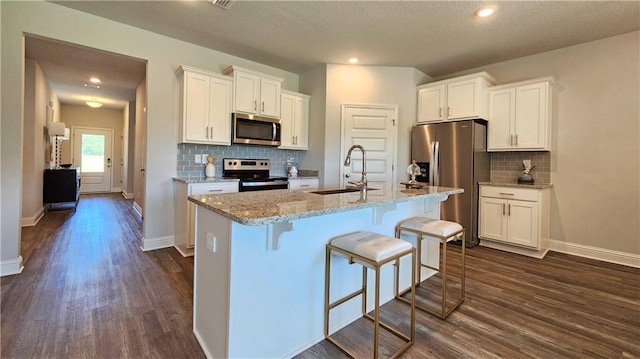 kitchen with appliances with stainless steel finishes, white cabinetry, light stone counters, an island with sink, and a kitchen bar