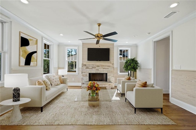 living room featuring a ceiling fan, wood finished floors, a stone fireplace, visible vents, and ornamental molding