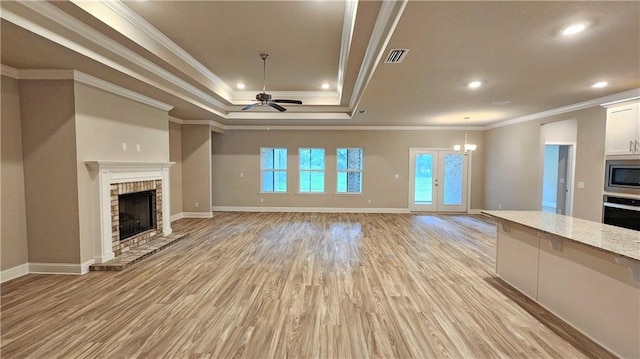 unfurnished living room with ceiling fan with notable chandelier, a raised ceiling, a brick fireplace, light wood-type flooring, and ornamental molding