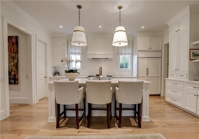 kitchen featuring light wood-type flooring, custom exhaust hood, a kitchen island with sink, paneled built in refrigerator, and pendant lighting