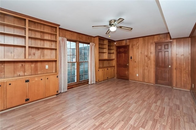 unfurnished living room with ceiling fan, wooden walls, ornamental molding, built in shelves, and light wood-type flooring