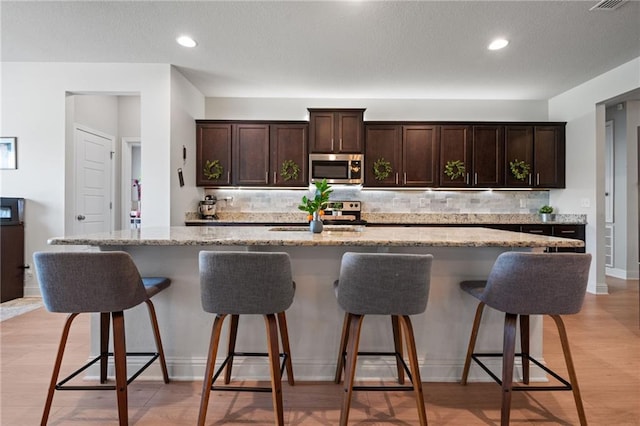 13077 Tara Point Dr, Mobile, AL 36695 - kitchen with appliances with stainless steel finishes, backsplash, dark brown cabinets, a kitchen breakfast bar, and a center island with sink