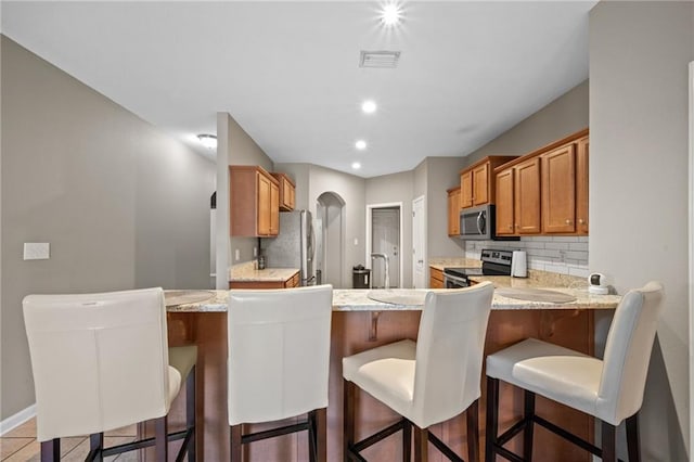 kitchen featuring stainless steel appliances, light stone counters, kitchen peninsula, a breakfast bar area, and decorative backsplash