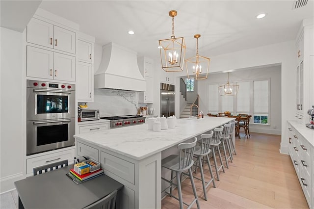 kitchen with premium range hood, white cabinetry, hanging light fixtures, a kitchen island with sink, and stainless steel appliances