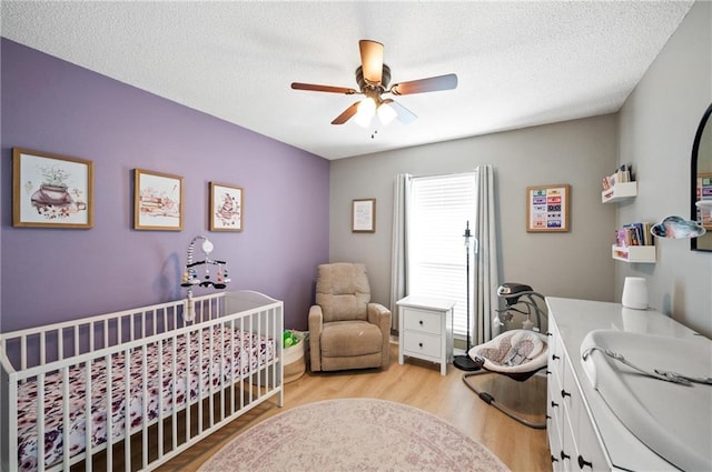 bedroom featuring ceiling fan, a nursery area, a textured ceiling, and light wood-type flooring