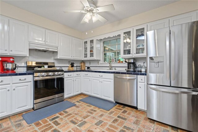 kitchen with tile counters, white cabinetry, under cabinet range hood, a sink, and appliances with stainless steel finishes