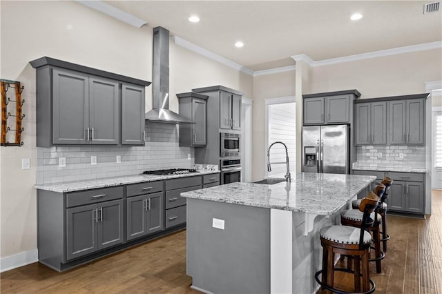 kitchen with a center island with sink, stainless steel appliances, gray cabinetry, wall chimney exhaust hood, and sink