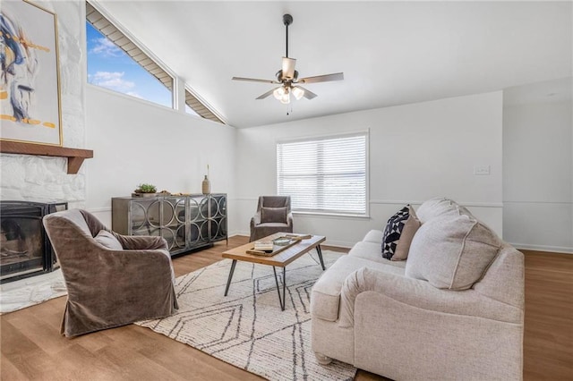 living room featuring wood finished floors, a stone fireplace, baseboards, ceiling fan, and vaulted ceiling