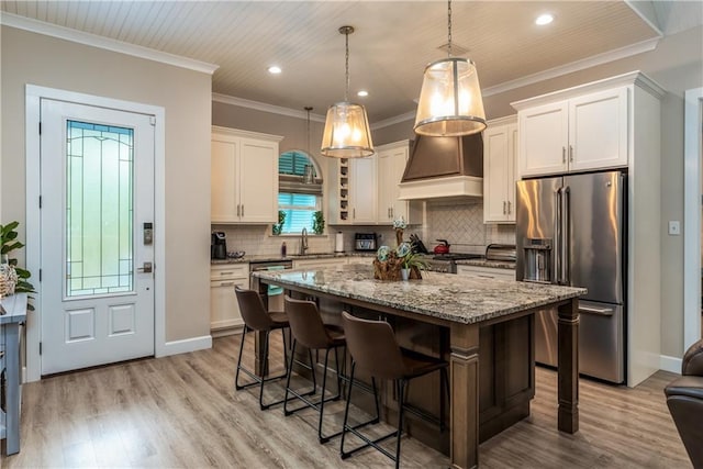 kitchen with appliances with stainless steel finishes, hanging light fixtures, white cabinets, a kitchen island, and custom exhaust hood