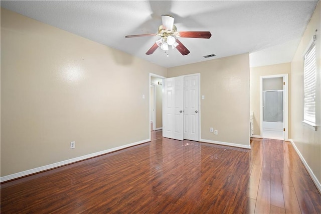 unfurnished bedroom with multiple windows, ceiling fan, ensuite bathroom, and dark wood-type flooring