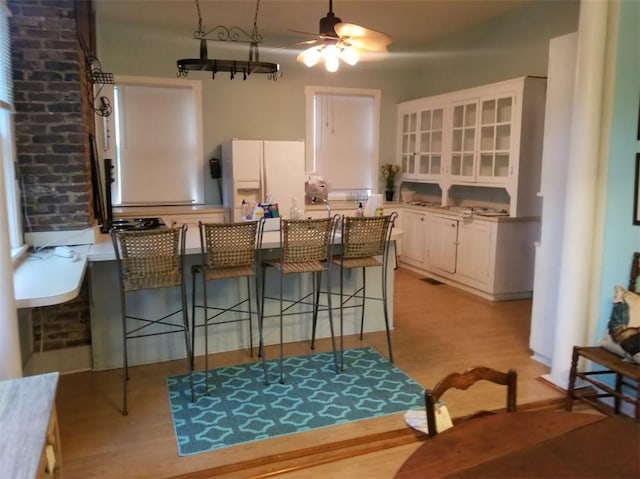 kitchen with ceiling fan, a breakfast bar area, white fridge with ice dispenser, white cabinetry, and light wood-type flooring