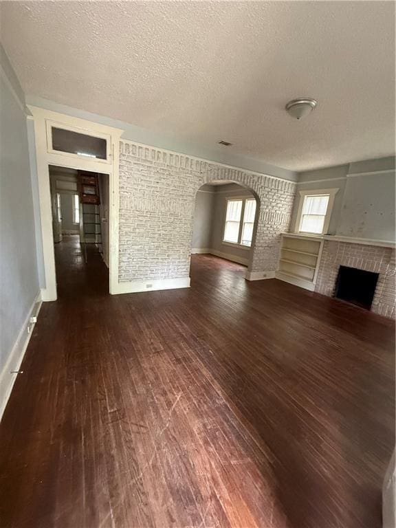unfurnished living room with a textured ceiling, arched walkways, dark wood finished floors, baseboards, and a brick fireplace